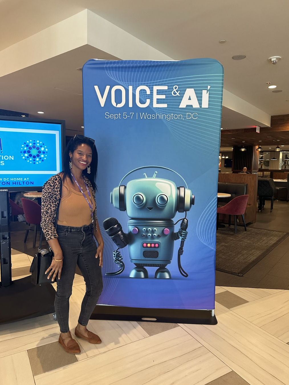 Jourdan standing in hotel lobby in front of a blue banner with text “Voice and AI” with robot holding a microphone.